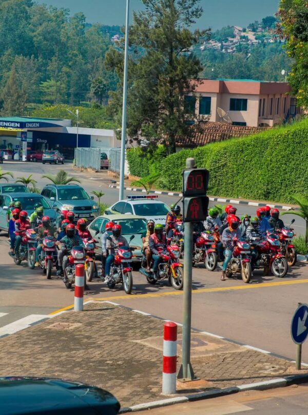 Motorcycle taxi riders and cars stopped at a pedestrian signal on a hilly city street